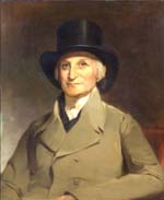 Seated white man with silver sideburns and top hat and wearing tan double breasted coat and white cravat.